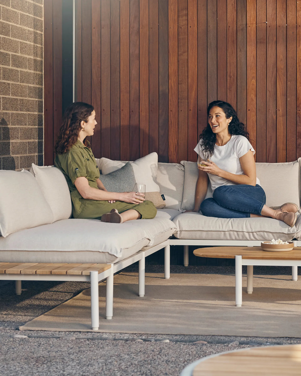 Two women talking while seated on an outdoor sectional with a teak end table