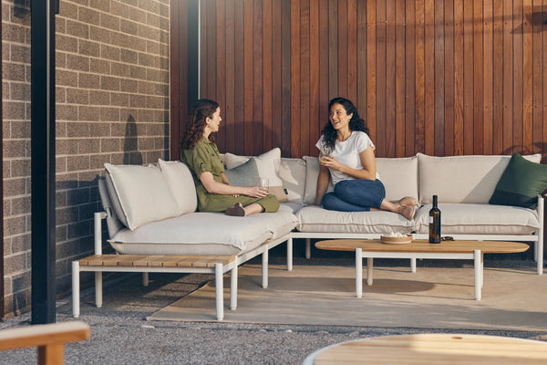 Two women talking while seated on an outdoor sectional with a teak end table