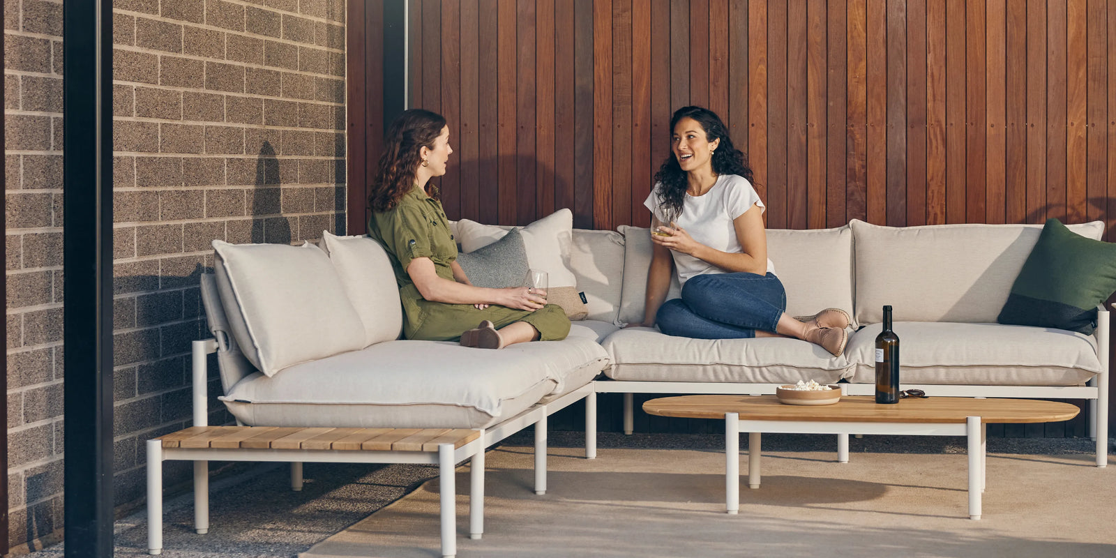 Two women talking while seated on an outdoor sectional with a teak end table