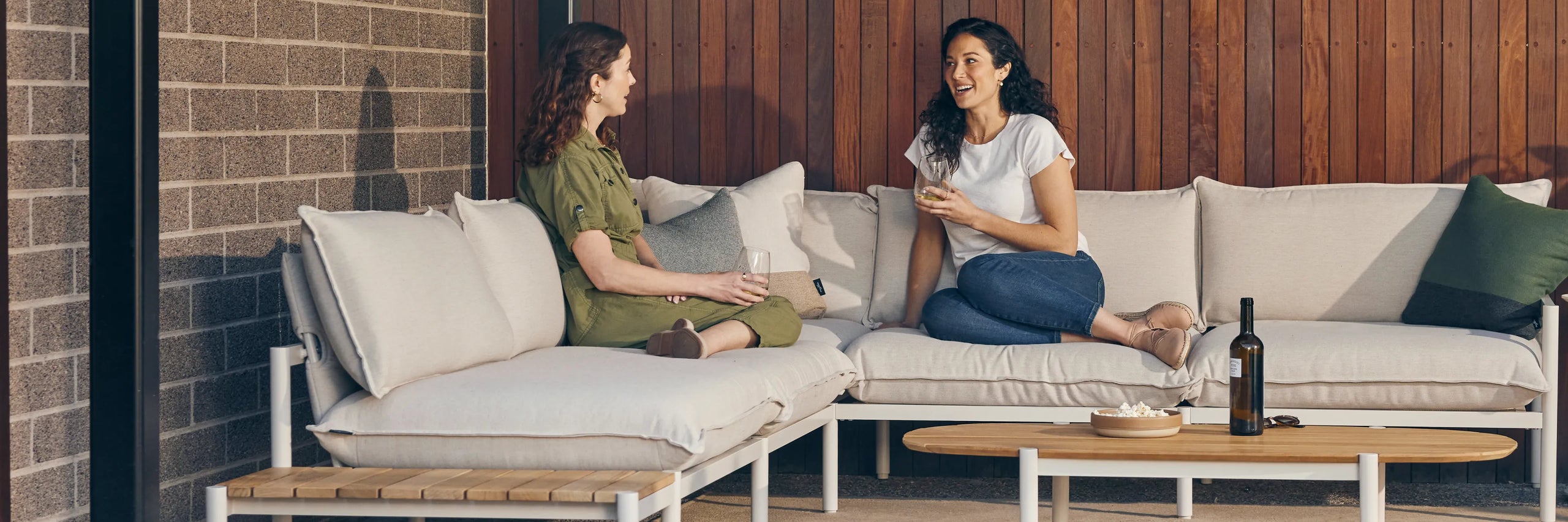 Two women talking while seated on an outdoor sectional with a teak end table