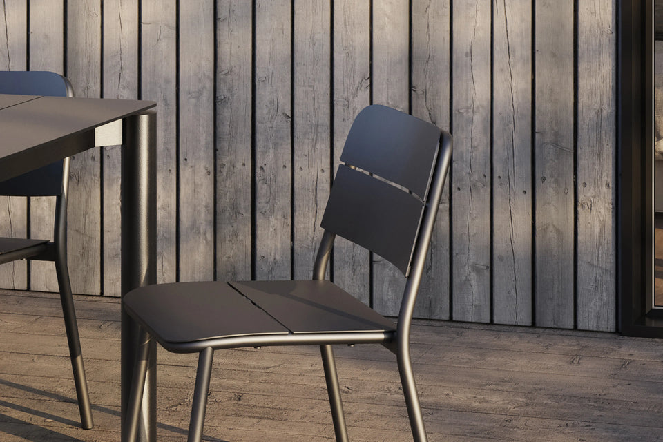 Neighbor Bandwidth Dining Chair in Charcoal beside matching table on a weathered wood deck with wood-slat wall backdrop