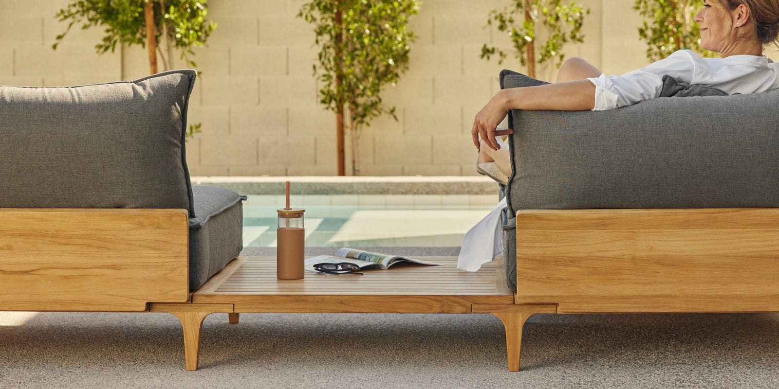Person seated on Neighbor Bluff Sofa with In-line Table in charcoal and teak on sunny patio, pool view