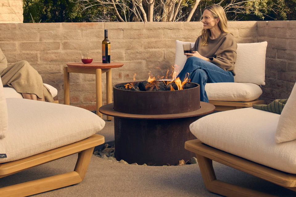 Two people relaxing in Neighbor Dweller Chairs with cream cushions and teak frame around a fire table on a walled patio at dusk