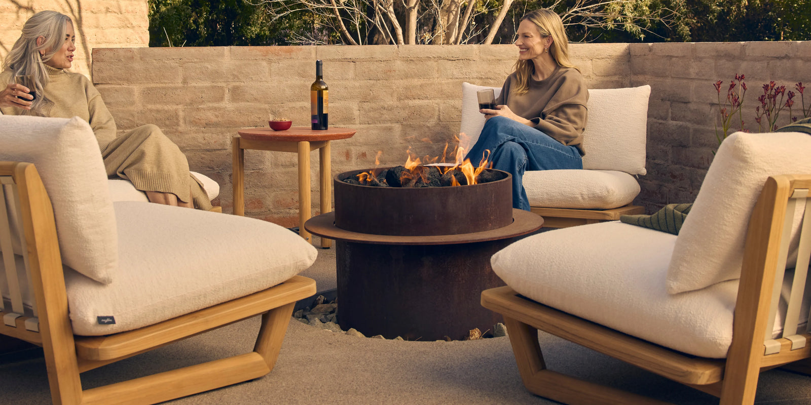Two people relaxing in Neighbor Dweller Chairs with cream cushions and teak frame around a fire table on a walled patio at dusk