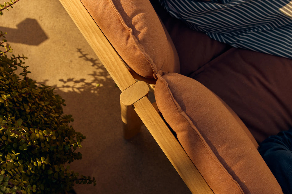 Overhead view of person on Neighbor Felloe Loveseat with rust cushion and teak frame on sunny patio near greenery