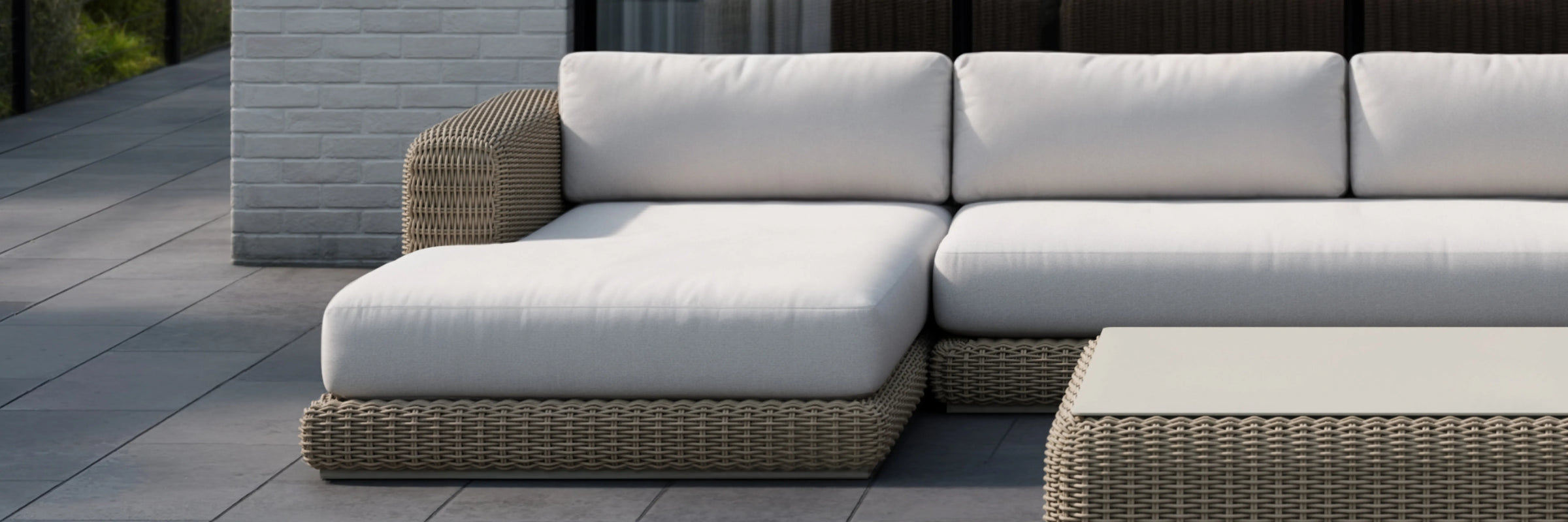 Neighbor Roundabout Chaise Sofa in dark wicker with charcoal cushions arranged on a paved patio with greenery