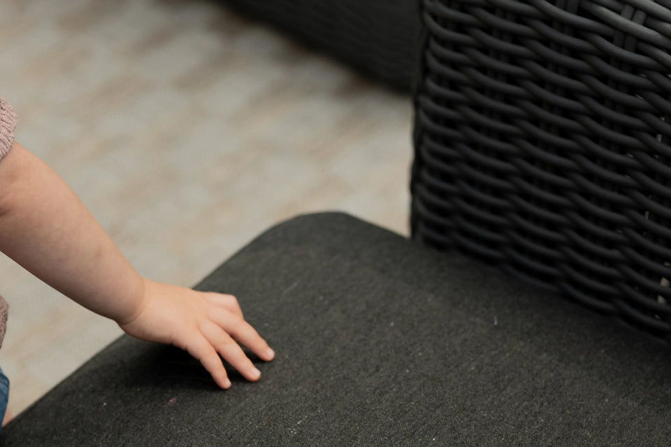 Child's hand resting on Neighbor Roundabout charcoal cushion, dark wicker arm panel visible beside