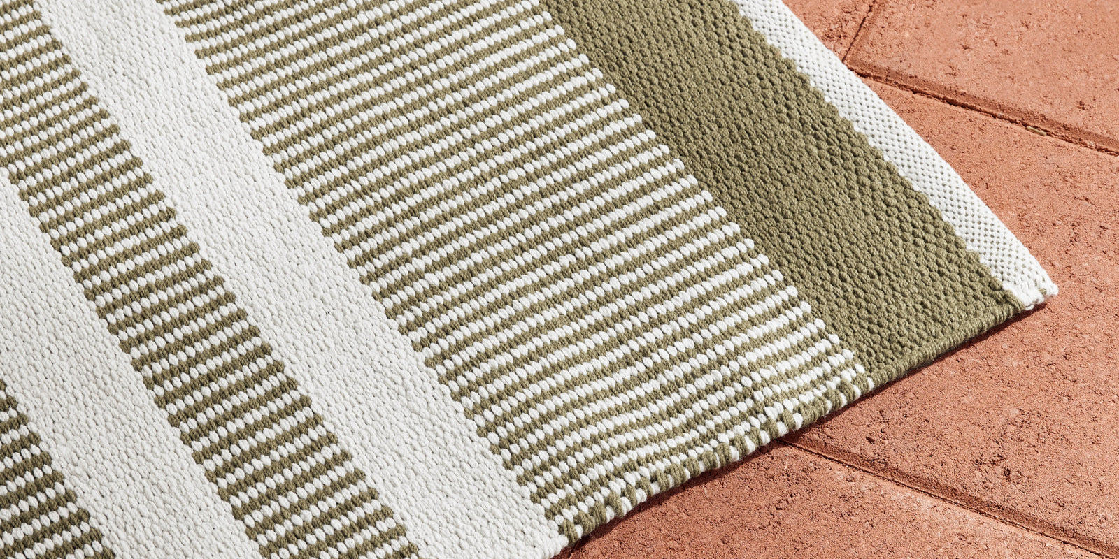 Neighbor Dune Outdoor Rug with green and white stripe pattern draped on terracotta tile floor, showing texture