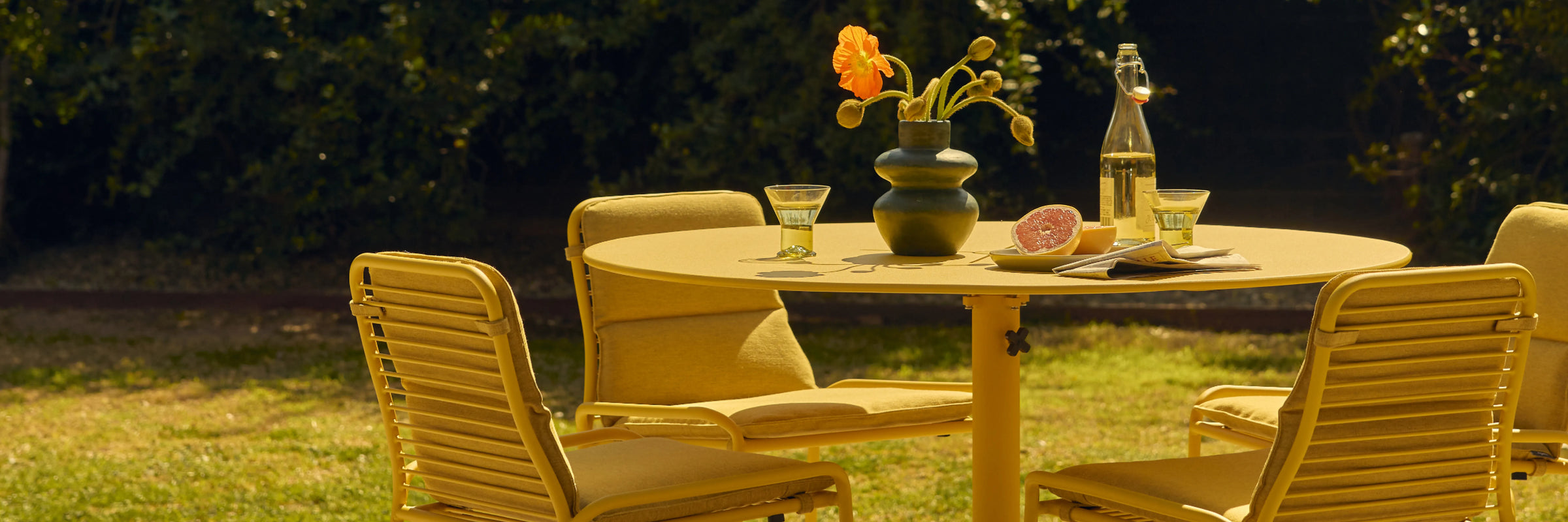 Neighbor XY Dining Set with round table and yellow aluminum chairs on a grass lawn at dusk, food and flowers on table