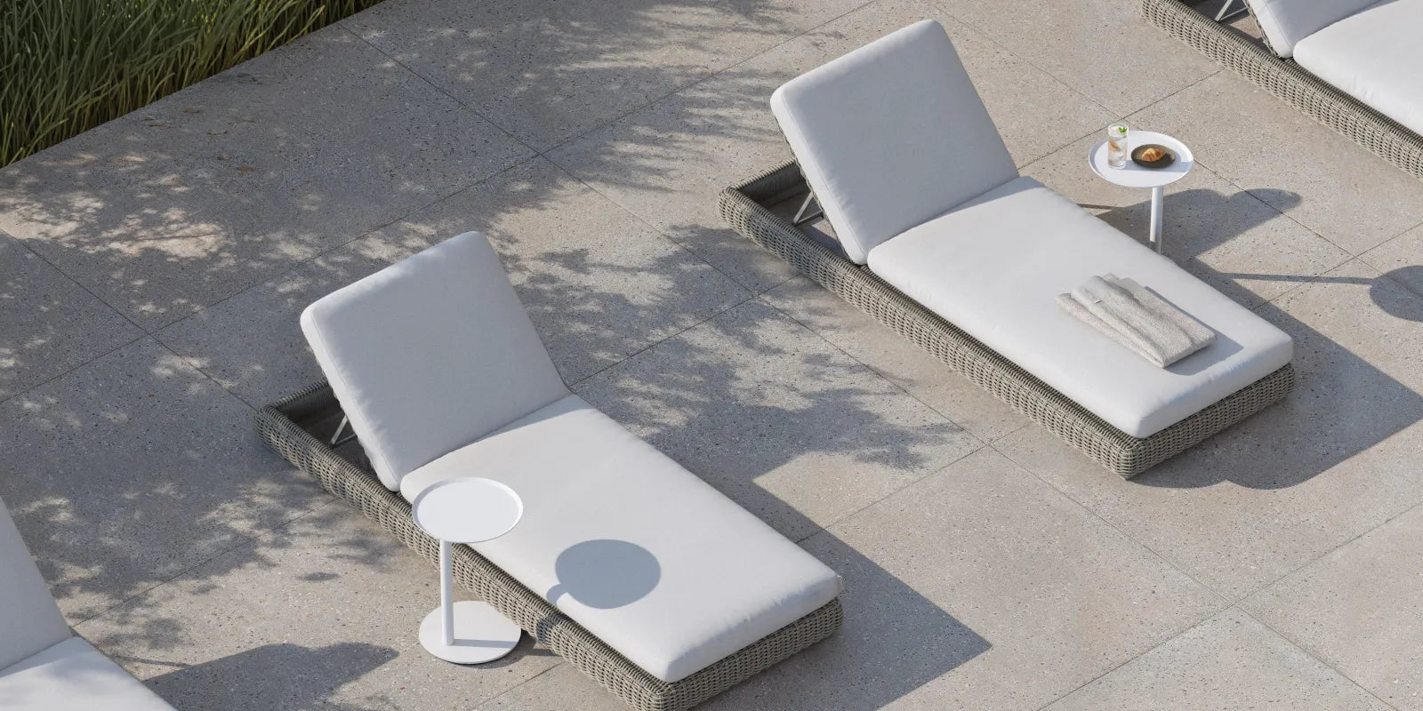 Top-down banner of four Neighbor Roundabout Sun Loungers in resin wicker with cream cushions arranged on a sunlit stone-tile patio, with a small round white side table holding a coffee and pastry between two of them; tall ornamental grasses cast soft shadows along the left edge.