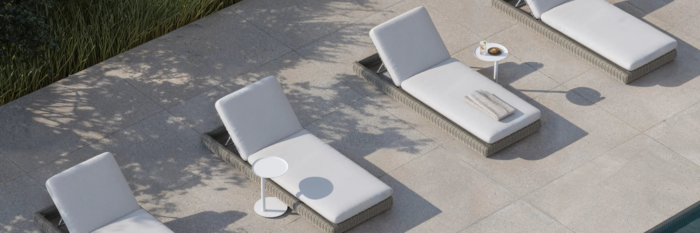 Top-down banner of four Neighbor Roundabout Sun Loungers in resin wicker with cream cushions arranged on a sunlit stone-tile patio, with a small round white side table holding a coffee and pastry between two of them; tall ornamental grasses cast soft shadows along the left edge.