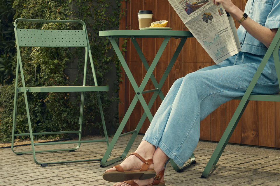 Banner of a woman in denim seated reading a newspaper at a sage-green Neighbor Take Two Bistro Set, folding bistro chair pulled up to the perforated round bistro table on brick pavers, with a stone planter wall, ornamental grasses, and a tall black ceramic planter framing the scene.