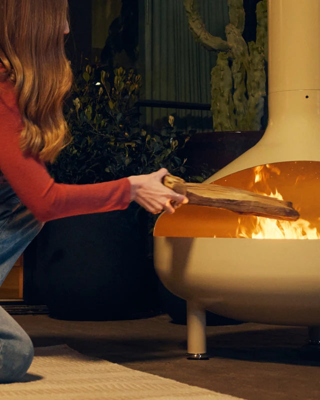 Wide banner of a woman in a red top kneeling to feed a log into the fire inside a cream-beige Neighbor Totem Chiminea on a covered patio at dusk, with warm pendant light, a woven-strap chair, and a stone-clad wall visible in the background.