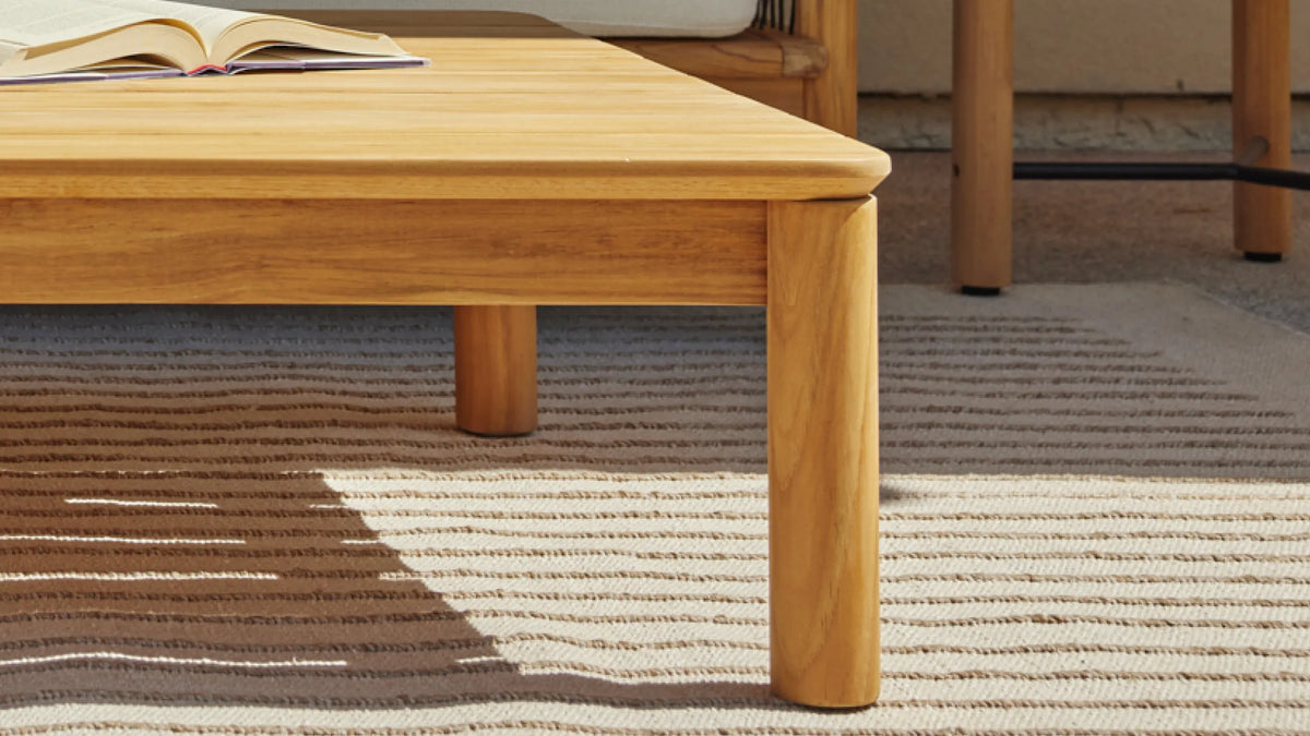 Teak tabletop edge and leg detail — front corner of Reel Coffee Table on a striped outdoor rug, open book on surface