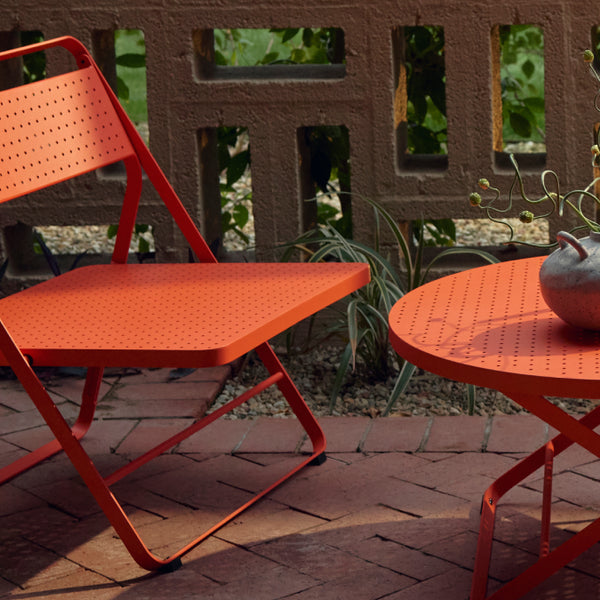 Three-quarter view of a coral-red Neighbor Take Two Lounge Chair beside a coral-red Take Two Coffee Table on a red-brick paver patio, with a candle, books and stoneware vase styled on the table; cinder-block lattice wall and tropical greenery behind.