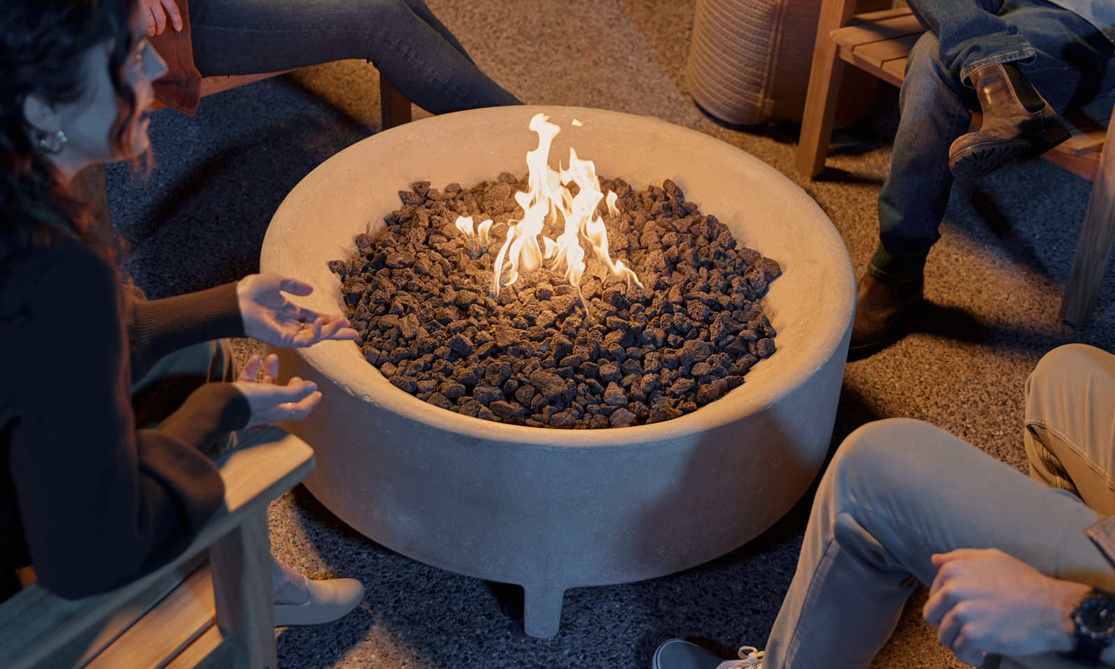 Group of people gathered around a round concrete fire table with lava rock and open flame at night — Fire Tables collection