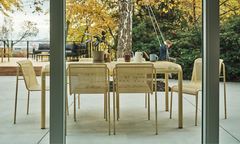 Perforated aluminum dining table and chairs in sand finish on a concrete terrace, viewed through open glass sliding doors with autumn foliage beyond — Outdoor Dining collection