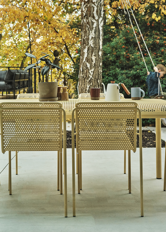 Perforated aluminum dining table and chairs in sand finish on a concrete terrace, viewed through open glass sliding doors with autumn foliage beyond — Outdoor Dining collection
