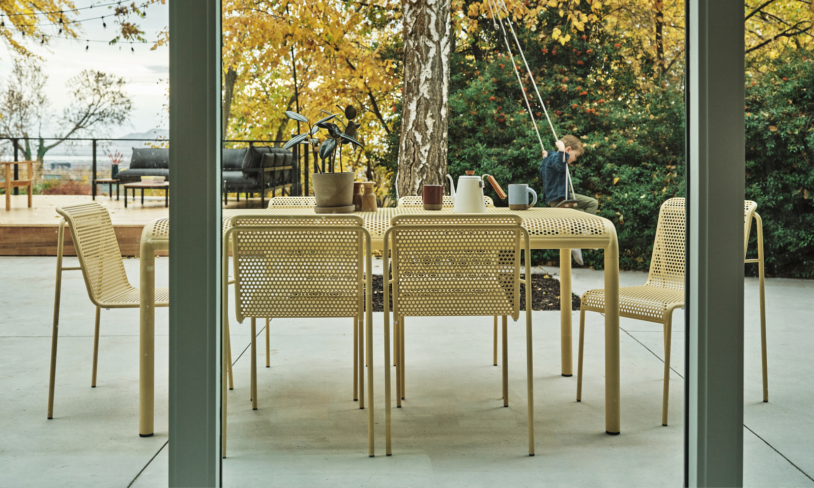 Perforated aluminum dining table and chairs in sand finish on a concrete terrace, viewed through open glass sliding doors with autumn foliage beyond — Outdoor Dining collection
