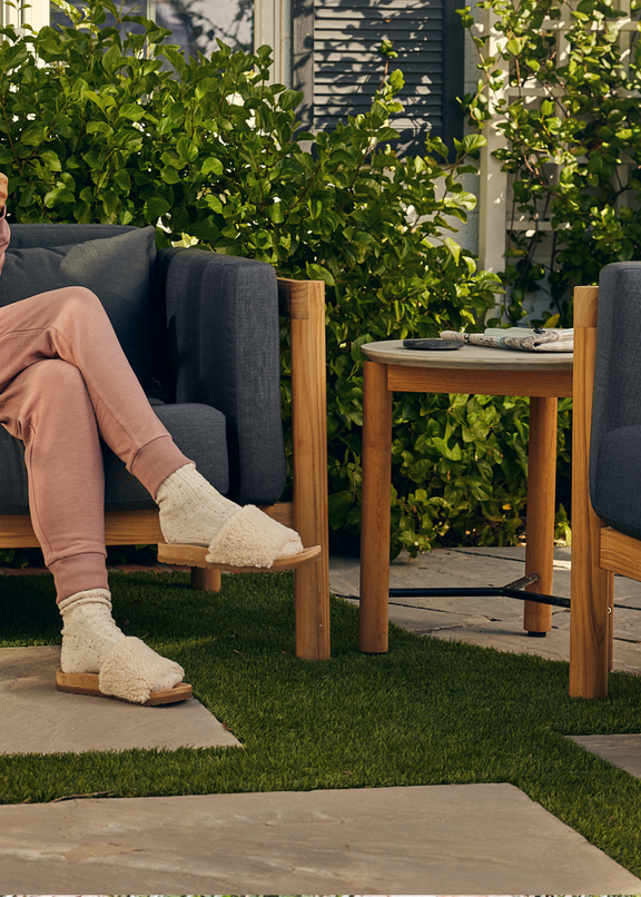 Woman in pink loungewear seated in a teak lounge chair with dark charcoal cushions holding a mug, with a second lounge chair and round side table in a lush garden — Outdoor Lounge collection