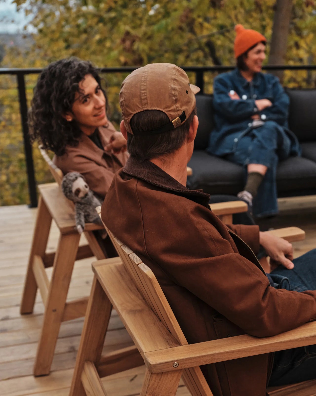 Adults and children gathered on teak outdoor chairs and a dark sectional sofa on a wood deck in autumn