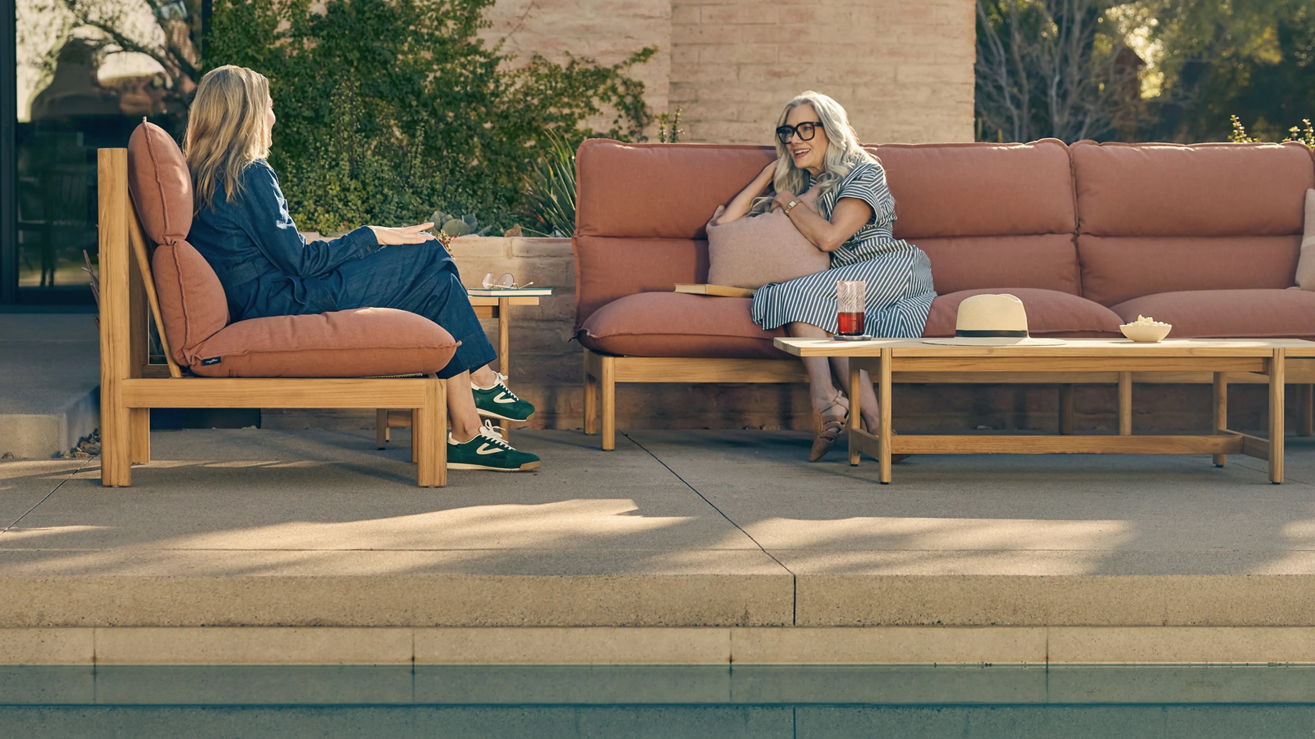 Two women relaxing poolside on teak outdoor sofa and lounge chair with terracotta cushions