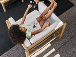 Person lounging on Neighbor Haven Chair with white cushion on concrete patio with tree shadows, holding a document