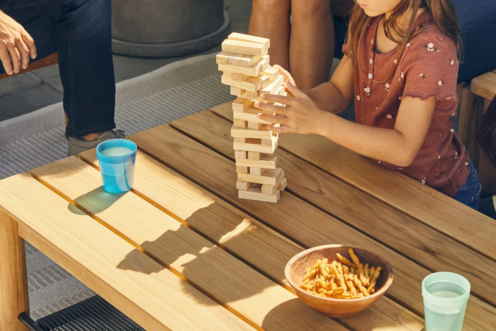 Neighbor Haven Square Coffee Table with concrete top in outdoor setting, with family playing Jenga, striped wood chairs nearby, sunny ambiance