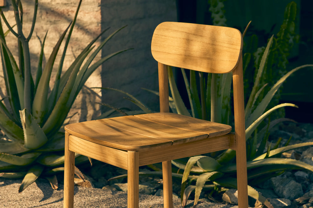 Neighbor Keeper Dining Chairs in natural teak with woven details in garden setting with potted plants and stone wall