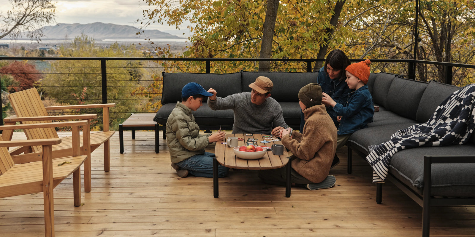 The Jones Family sitting around their Terra teak coffee table, playing games with the Terra Sectional and fall leaves in the background.