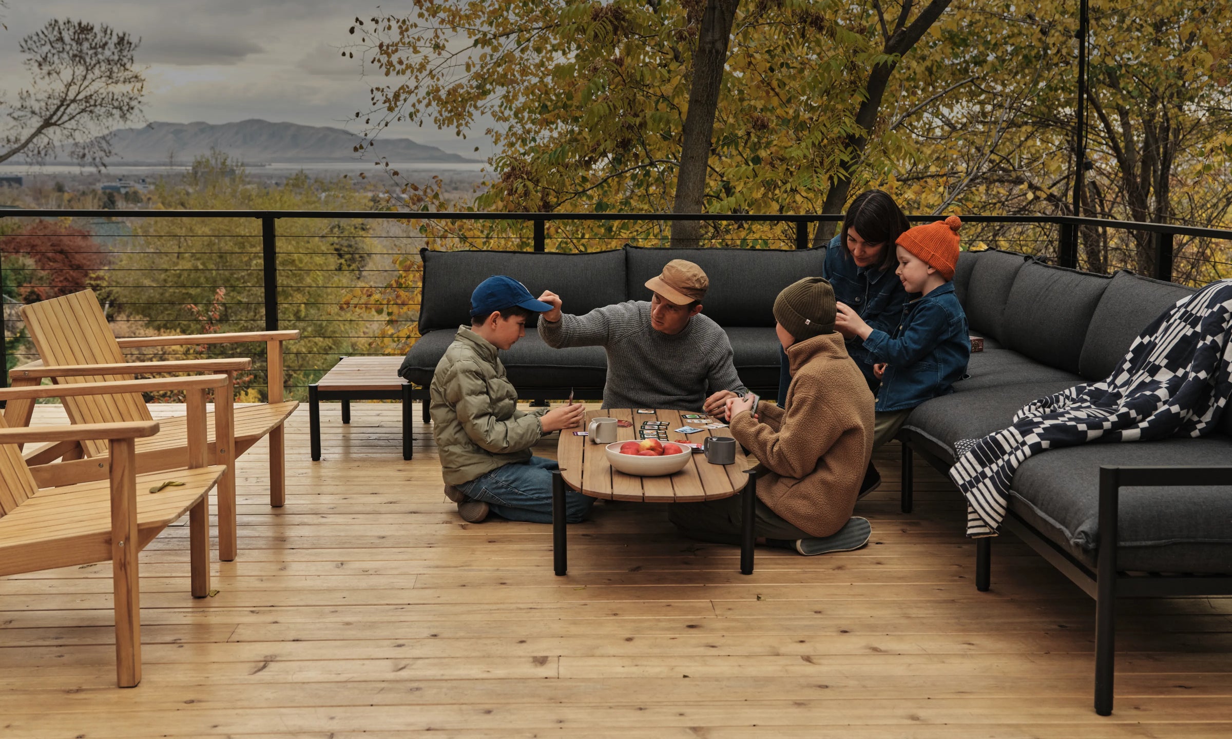 The Jones family—Cree and Amanda with three of their children, sitting atop their dark gray outdoor sectional on their wooden deck, playing cards on the teak coffee table. Mountains and fall foliage are in the background.