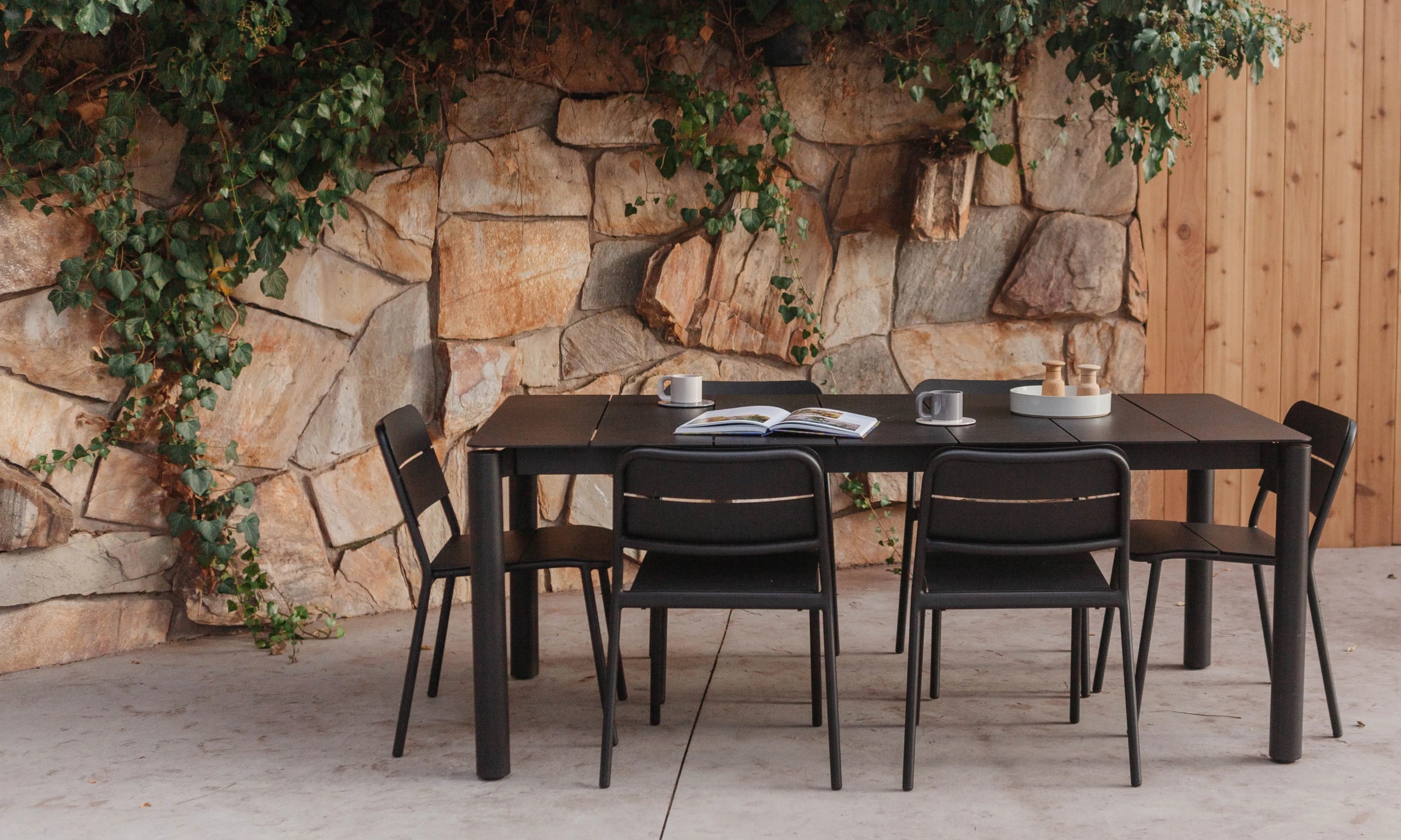 A rectangular black outdoor dining table with six matching black chairs, styled on a patio in front of a rustic stone wall with climbing green ivy and a wooden fence.