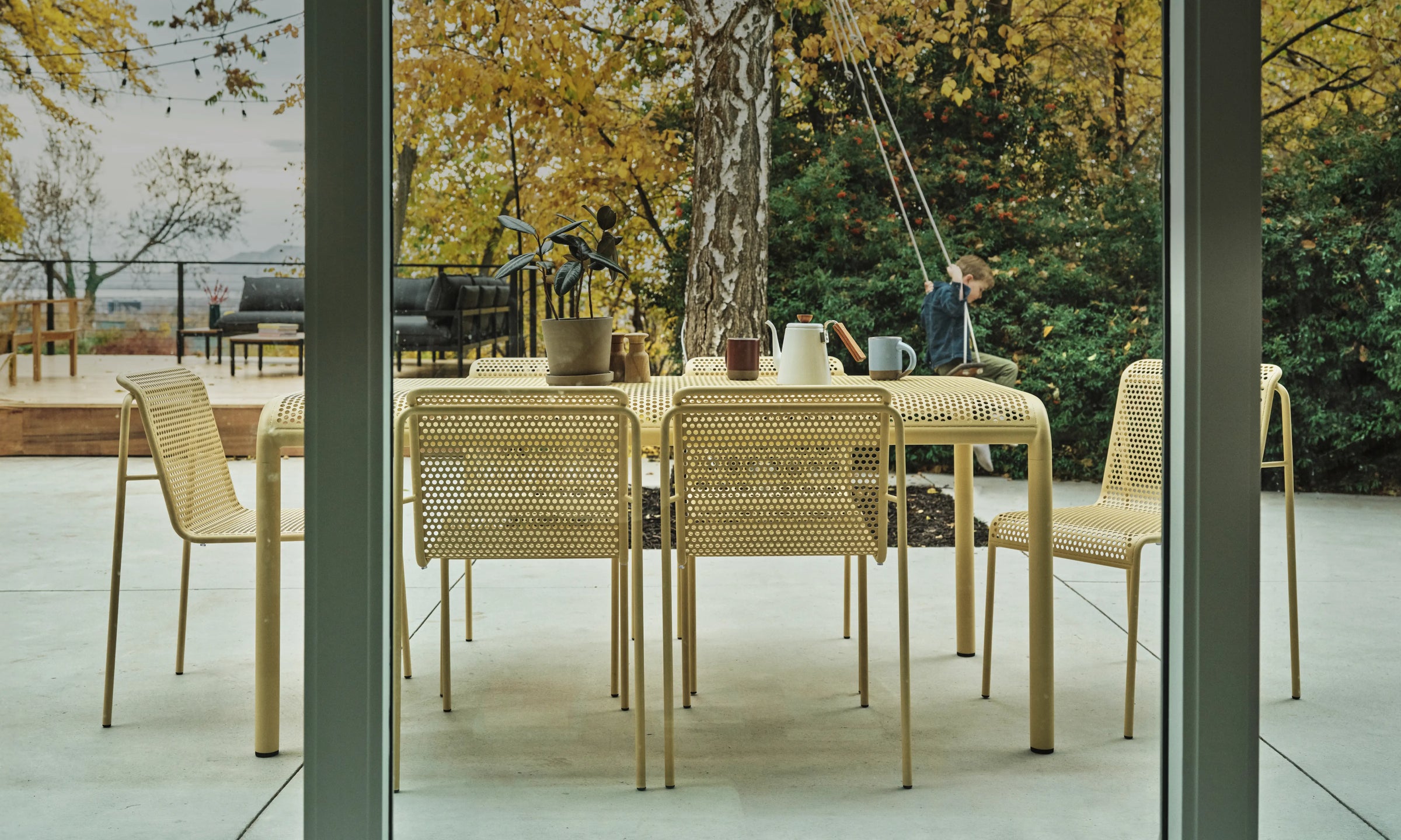 A modern outdoor dining set consisting of a rectangular perforated table and six perforated chairs, shown on a patio with a child sitting on a swing in the background.