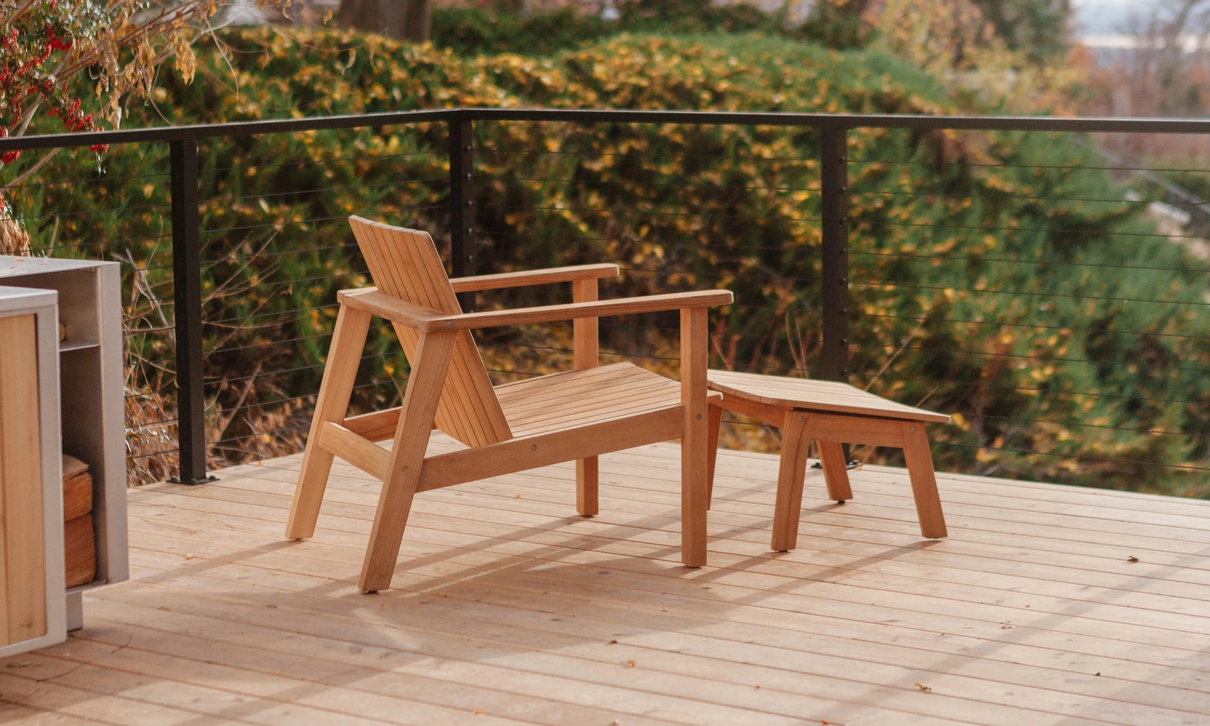 A teak wood outdoor lounge chair with a matching small side table on a wooden deck, with a black railing and green foliage in the background.