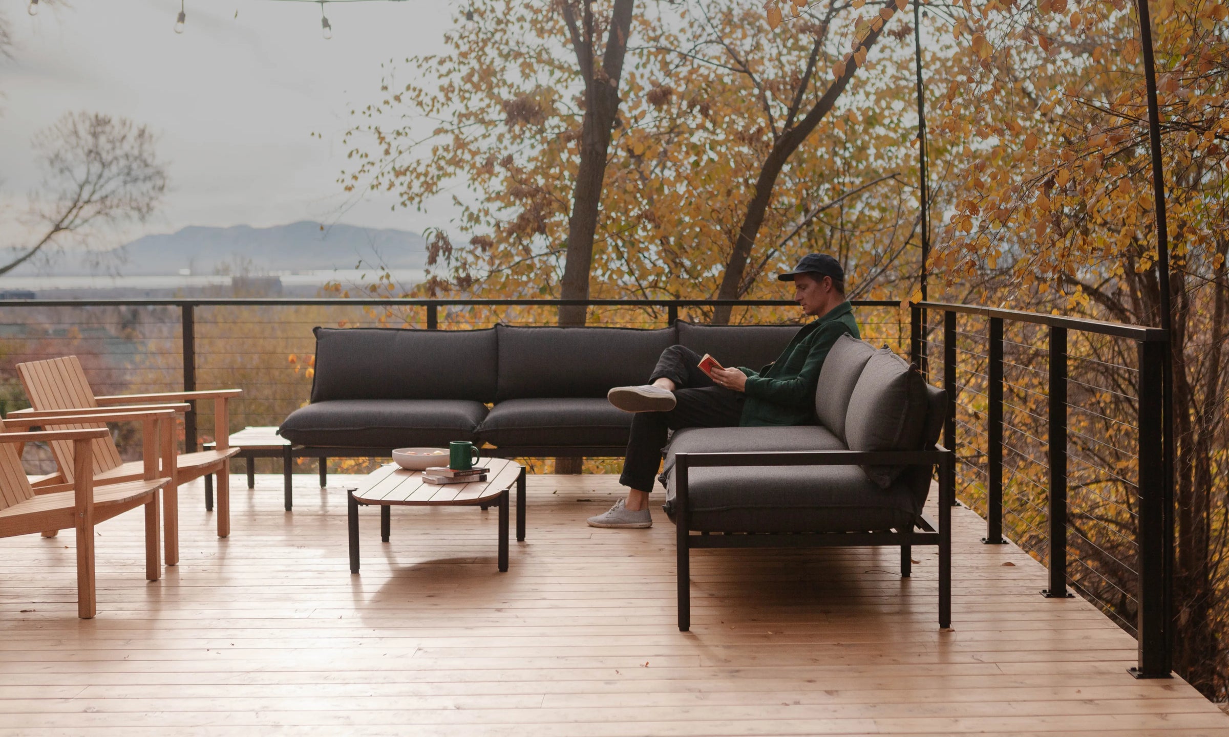 A man sits on a dark gray outdoor sectional sofa on a wooden deck, holding a red cup. The background features a scenic view of trees with yellow autumn leaves.