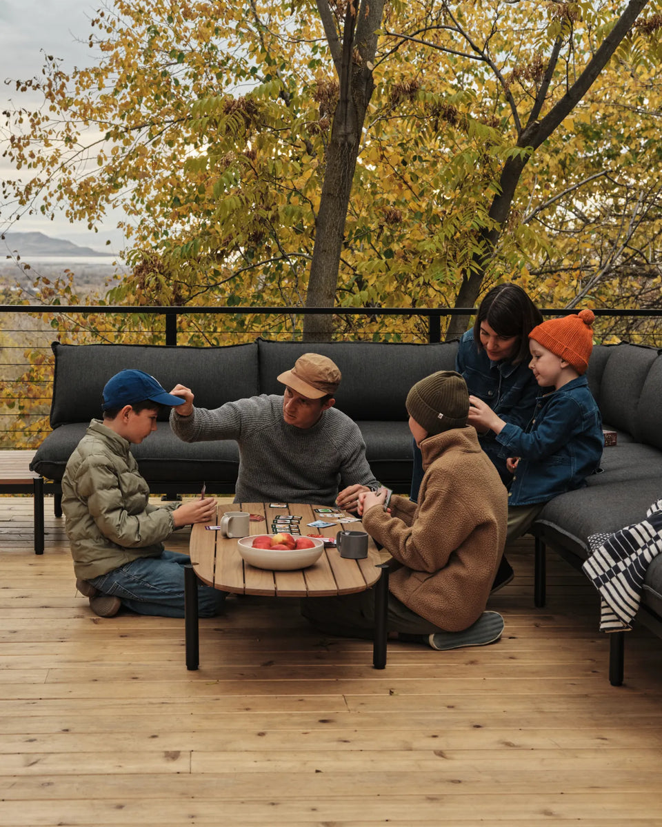 The Jones Family sitting around their Terra teak coffee table, playing games with the Terra Sectional and fall leaves in the background.