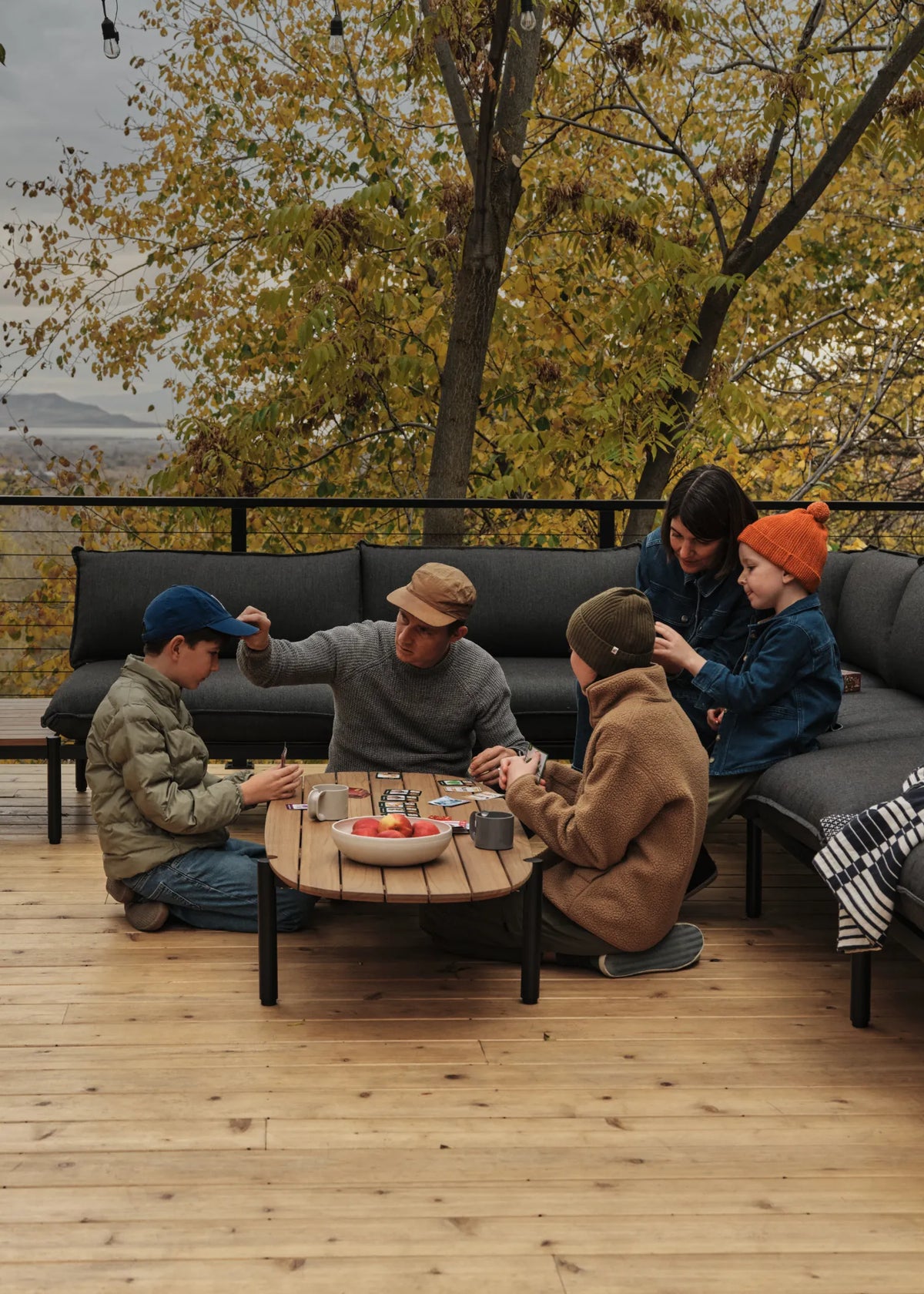 The Jones family—Cree and Amanda with three of their children, sitting atop their dark gray outdoor sectional on their wooden deck, playing cards on the teak coffee table. Mountains and fall foliage are in the background.