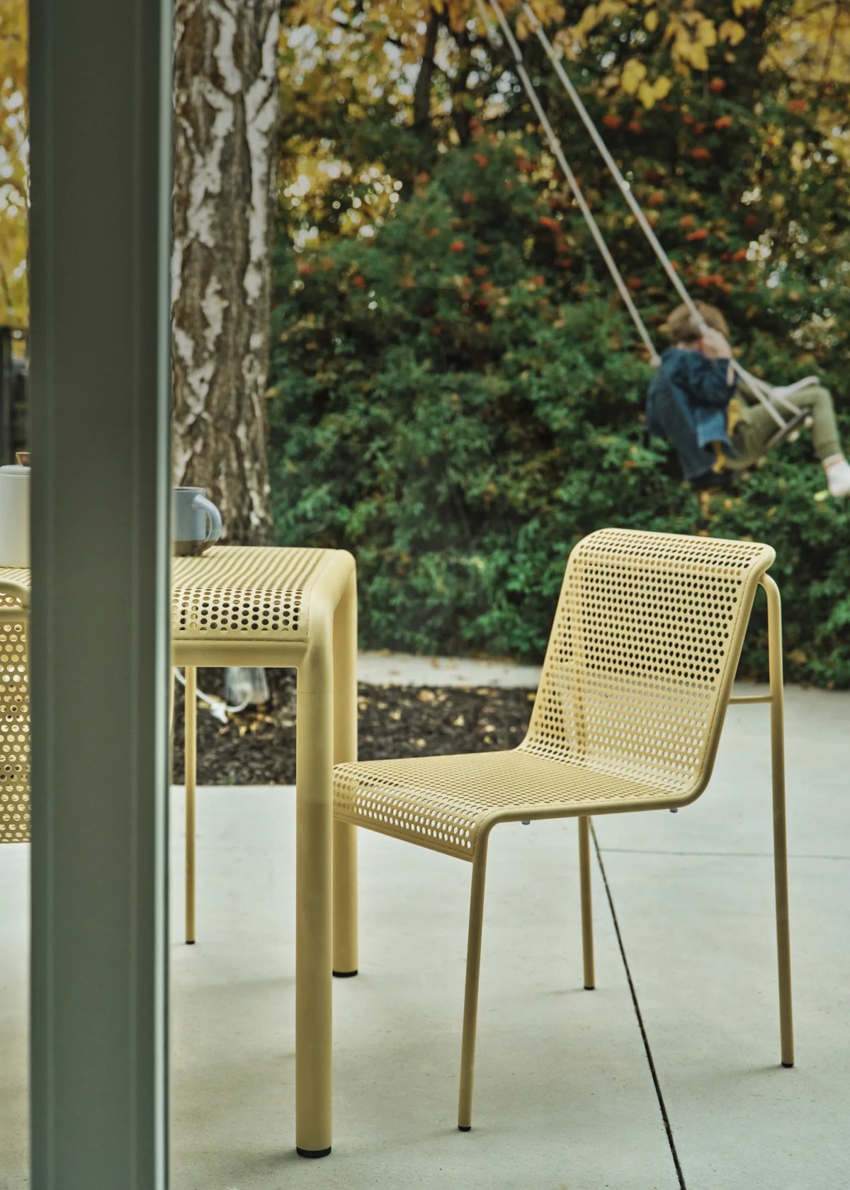 A cropped modern outdoor dining set consisting of a rectangular perforated table and a perforated chair, shown on a patio with a child sitting on a swing in the background.