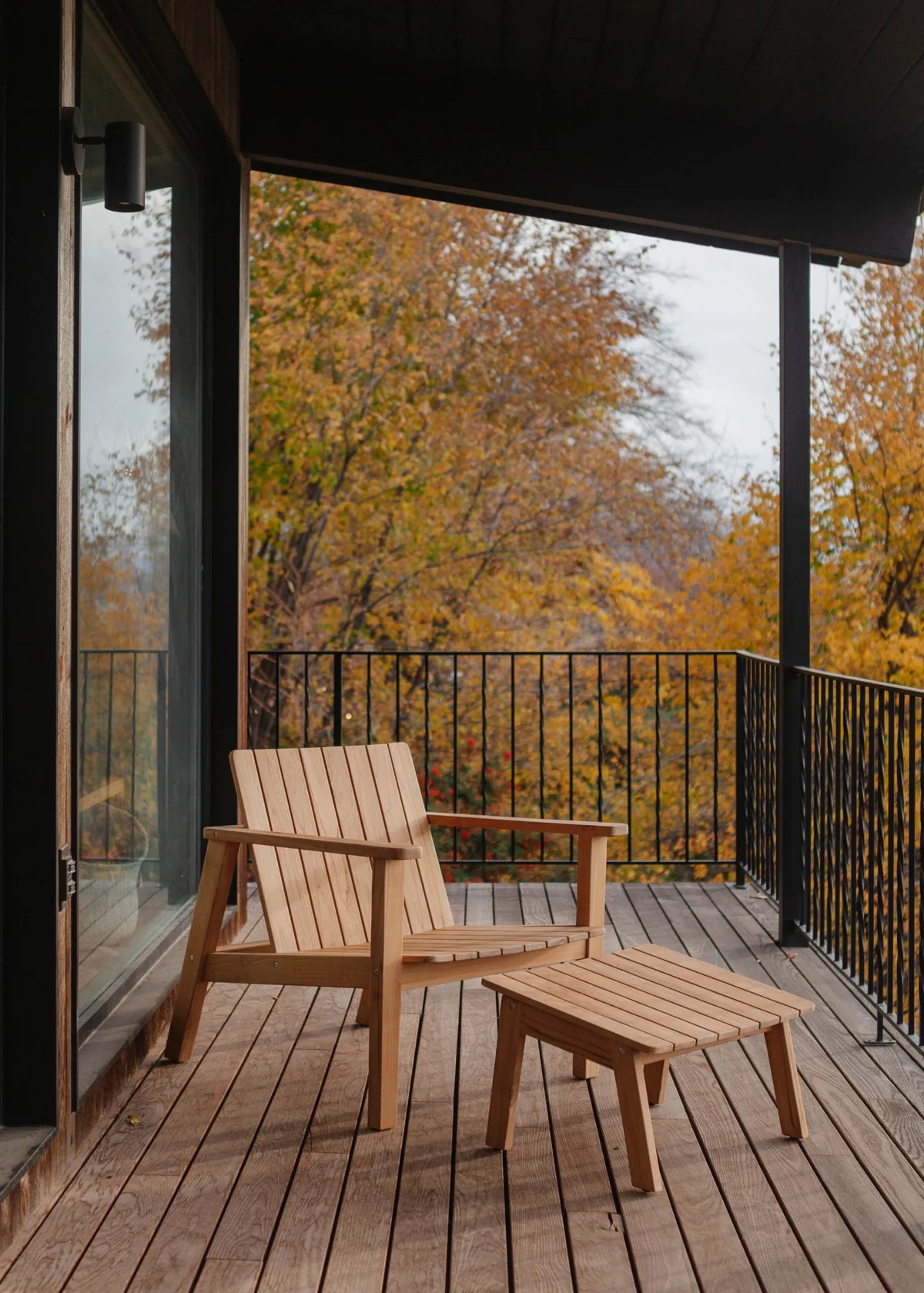 A wooden Adirondack-style chair and matching ottoman on a deck overlooking a fall forest with yellow and orange trees.