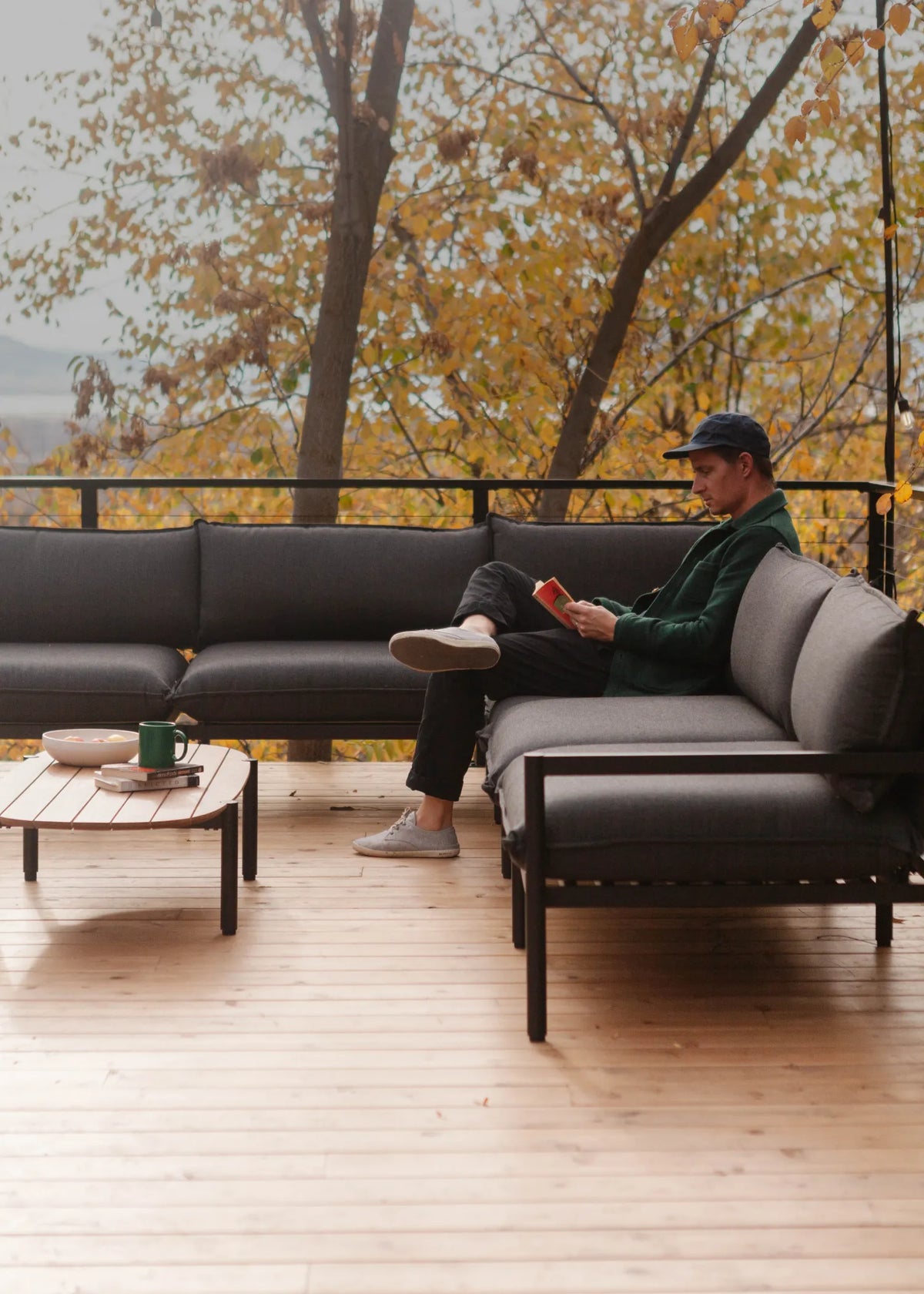 A man sits on a dark gray outdoor sectional sofa on a wooden deck, holding a red cup. The background features a scenic view of trees with yellow autumn leaves.