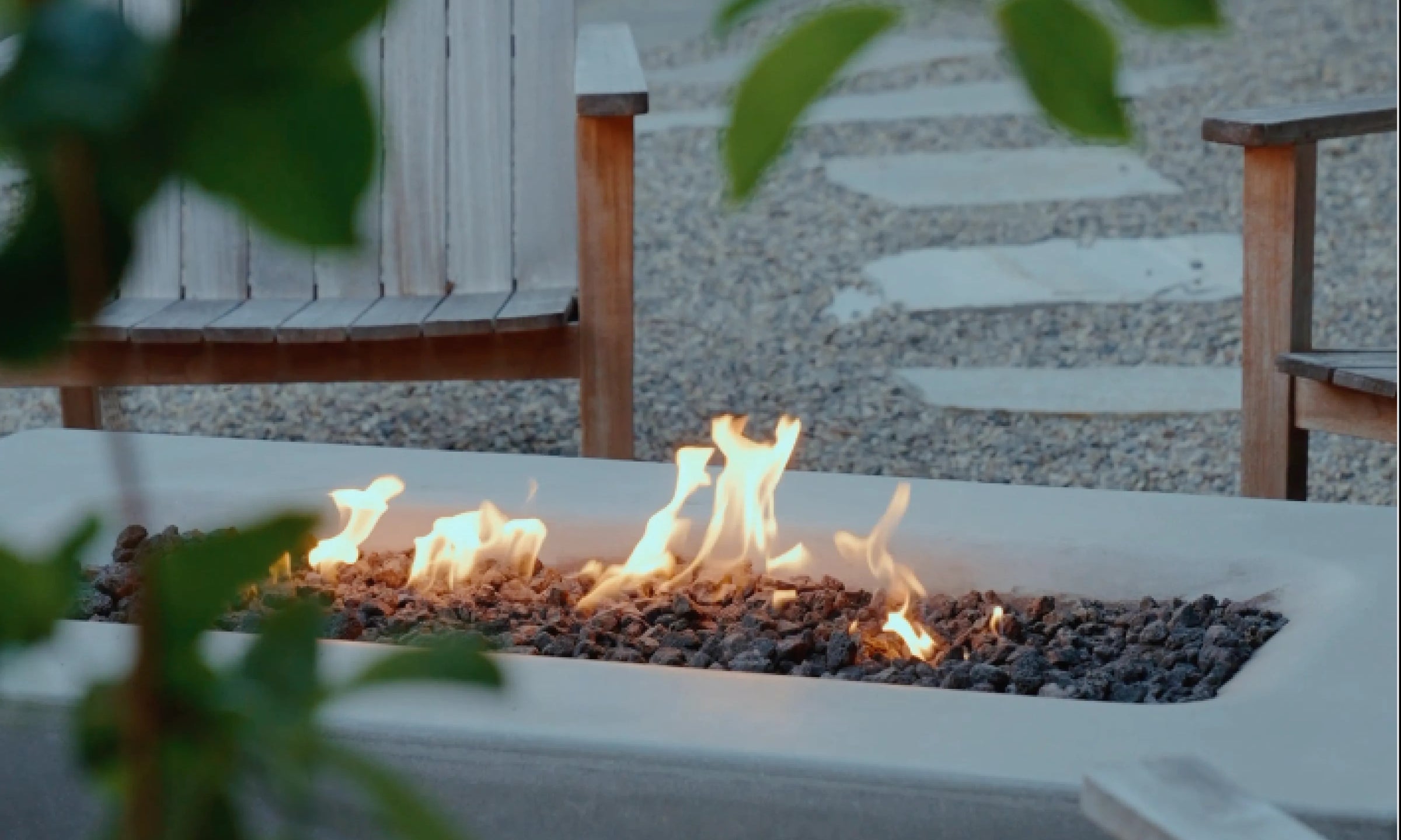 Outdoor rectangular fire pit with dark gravel and visible flames, set in a patio or garden area with wooden chairs and a stone path in the background.