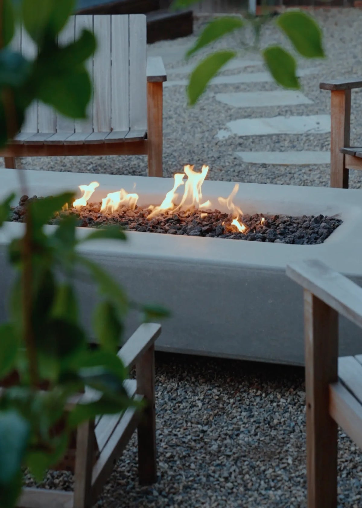A rectangular outdoor fire pit with a white concrete base, filled with dark gravel and active flames, situated in a patio setting with wooden chairs and a stone path.