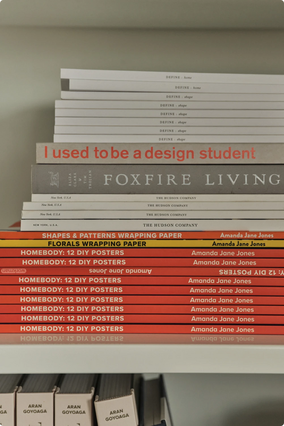 A stack of books and wrapping paper designs by Amanda Jane Jones, including Foxfire Living and I Used to Be a Design Student