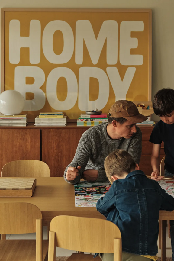 The Jones family gathers around their dining table coloring together, with a yellow "Homebody" print hanging on the wall behind them
