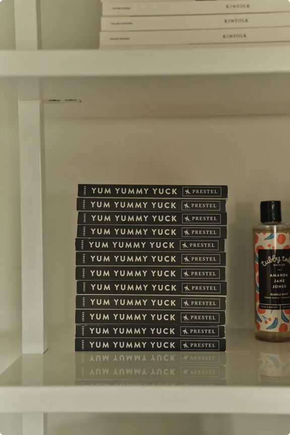 A stack of Amanda Jane Jones's "Yum Yummy Yuck" books on a shelf beside a bottle of Tubby Todd bubble bath and stacked Kinfolk magazines