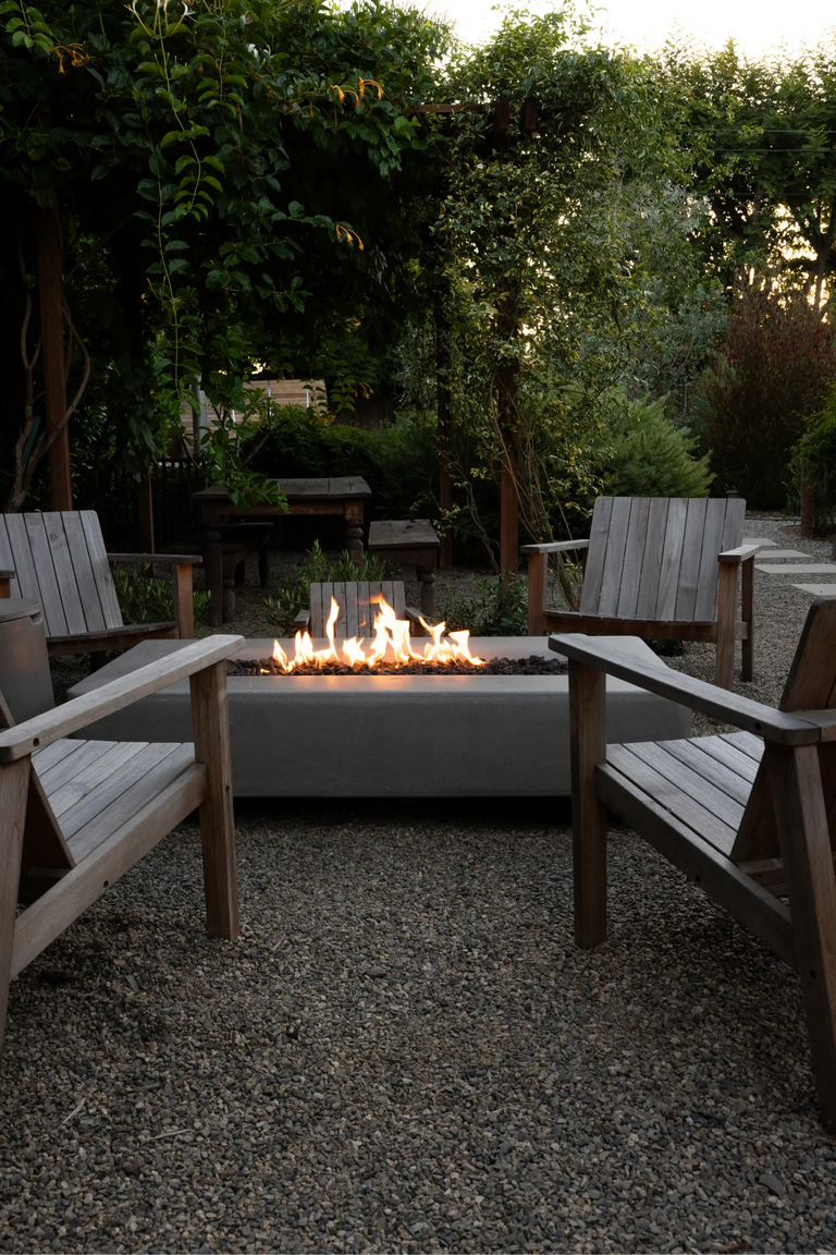 A lit Rook Fire Table surrounded by wooden Adirondack chairs in a lush, tree-lined backyard
