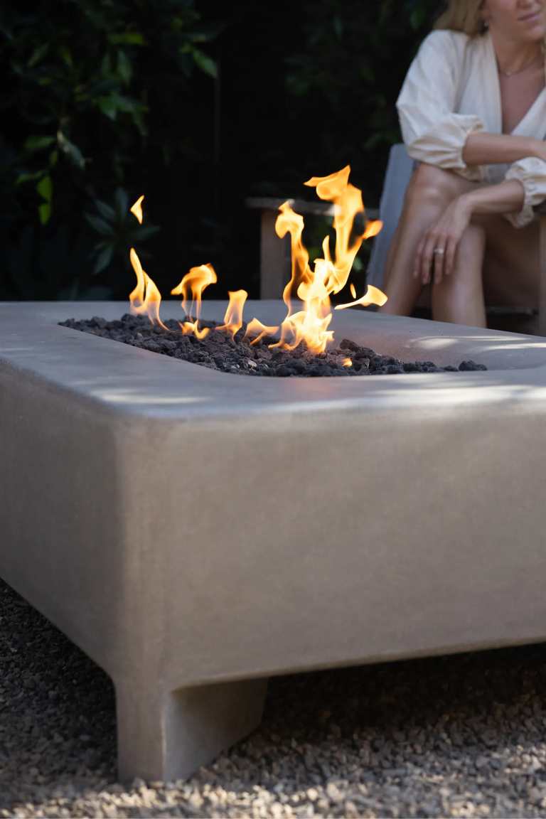 Close-up of a Rook Fire Table burning at dusk with wooden chairs and a seated figure in the background