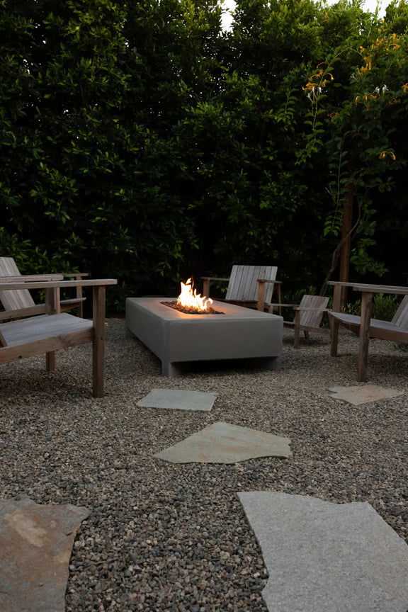 Wide view of a lit Rook Fire Table surrounded by wooden chairs in a dusk backyard setting
