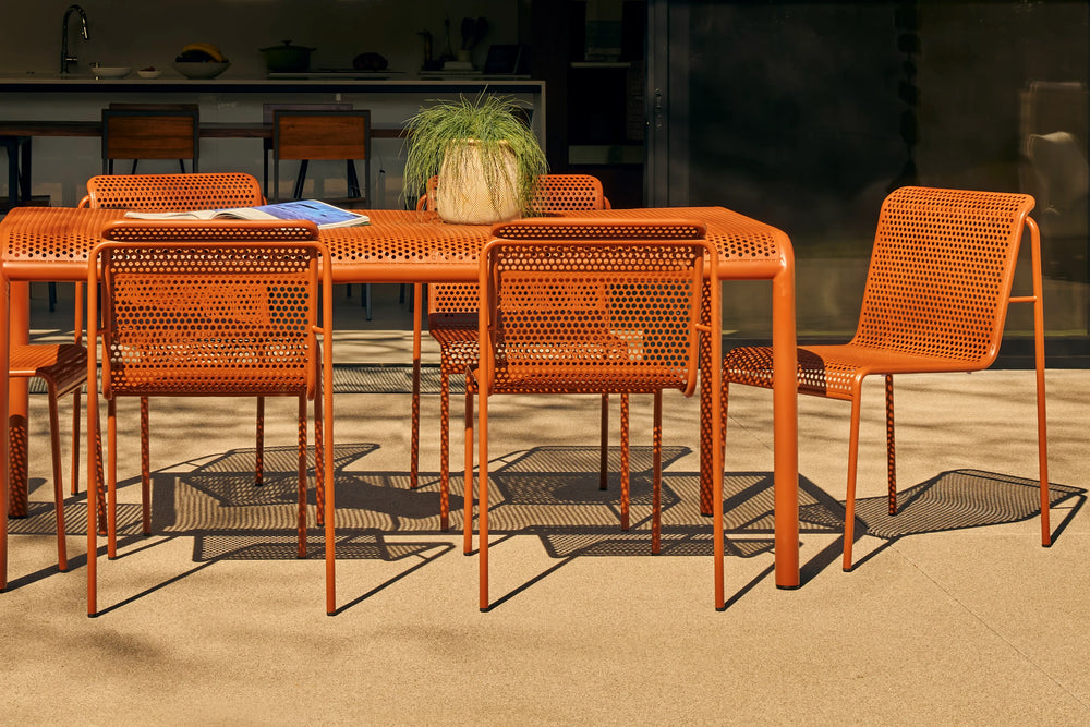 Neighbor Perf Dining Table with orange perforated metal chairs arranged on sand surface with tropical plants and glass walls