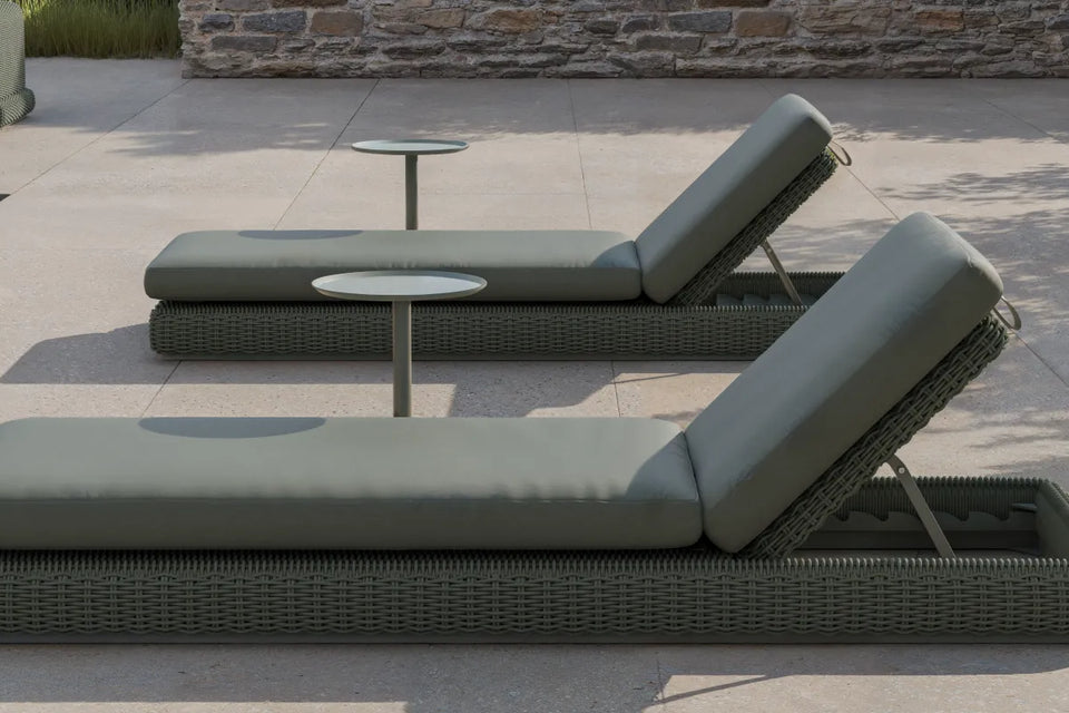 Wide poolside banner of two Neighbor Roundabout Sun Loungers in dark olive-gray resin wicker with matching olive-gray cushions arranged side-by-side on a sunlit stone-tile pool deck, with a small white round side table between them, ornamental grasses to the right, and a stacked-stone wall in the background.